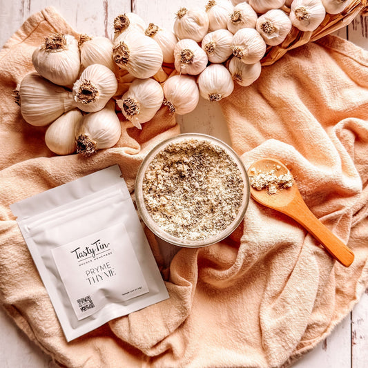 Tasty Tin Pryme Thyme seasoning blend showing natural clumping, surrounded by garlic bulbs on a peach towel — all-natural, filler-free seasoning