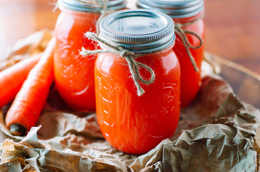 Carrot apple juice in mason jars with twine tide around the side carrots sitting beside them and everything on a crinkly brown paper bag