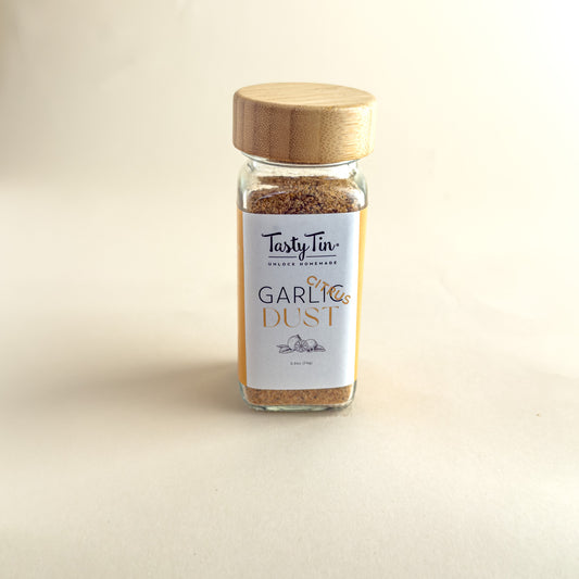 A glass spice jar of TastyTin Garlic Dust with a light wood lid, set against a plain beige background.