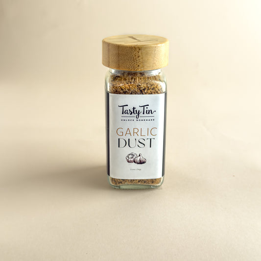 A small glass jar of TastyTin Garlic Dust seasoning with a light wood lid, centered on a warm beige background.