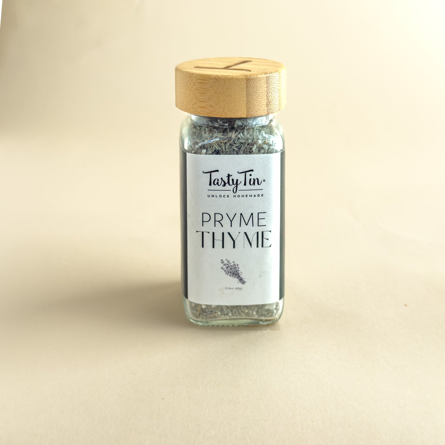 A small glass jar of TastyTin Pryme Thyme seasoning with a light wood lid, set against a plain beige background.