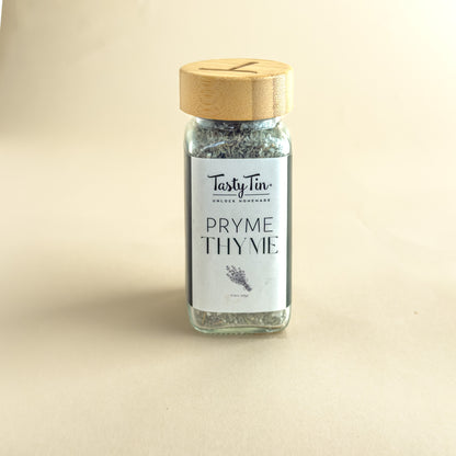 A small glass jar of TastyTin Pryme Thyme seasoning with a light wood lid, set against a plain beige background.