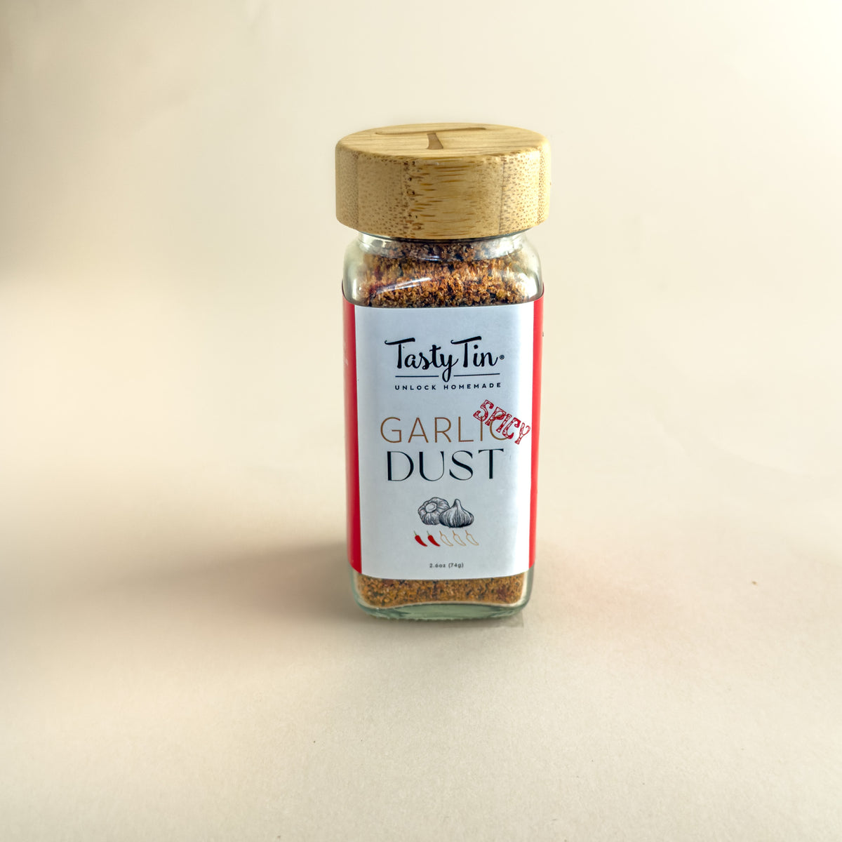 A glass jar of TastyTin Garlic Dust seasoning with a light wood lid, set against a plain beige background.