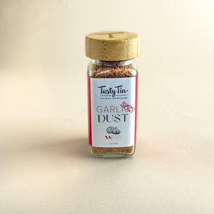 A glass jar of TastyTin Garlic Dust seasoning with a light wood lid, set against a plain beige background.
