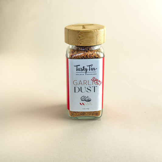 A glass jar of TastyTin Garlic Dust seasoning with a light wood lid, set against a plain beige background.