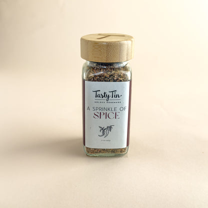 A small glass spice jar with a light wood lid and a white label reading 'TastyTin' and 'A Sprinkle of Spice', centered on a warm beige background.