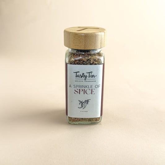 A small glass spice jar with a light wood lid and a white label reading 'TastyTin' and 'A Sprinkle of Spice', centered on a warm beige background.