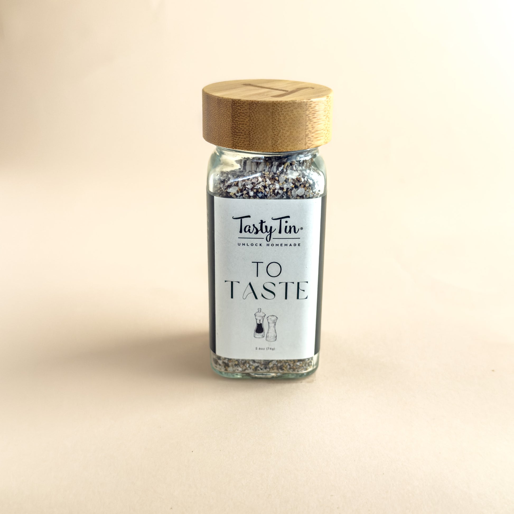 A small glass spice jar with a light wood lid and a white label reading 'TastyTin' and 'TO TASTE', filled with dark dried herbs, centered on a warm beige background.