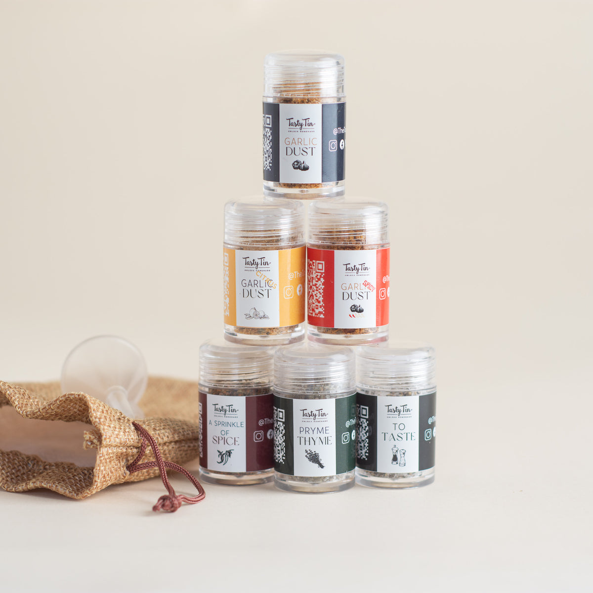 Six small clear spice jars arranged in a pyramid on a light beige background, with a burlap sack and scoop beside them. The labels show different spice flavors including garlic dust, rose dust, cumin dust, curry powder, saffron thyme, and to taste.