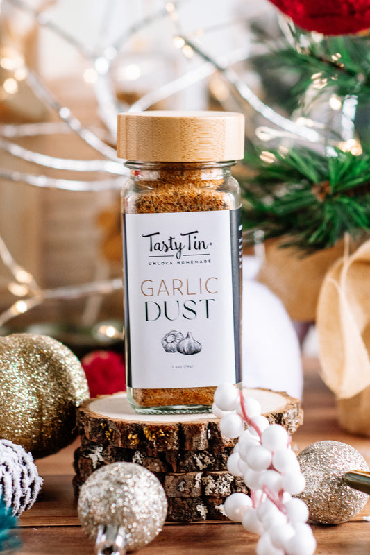 Tasty Tin garlic dust seasoning shaker with wooden lid displayed on wood slices surrounded by holiday decorations
