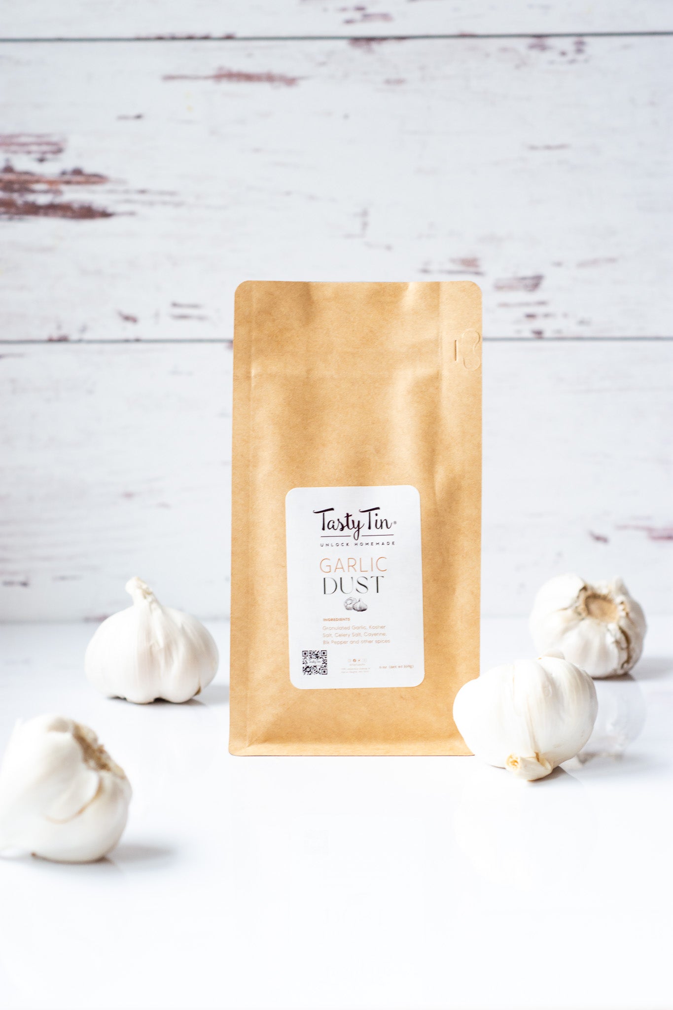 Tasty Tin garlic seasoning refill pouch standing on white surface with fresh garlic bulbs around