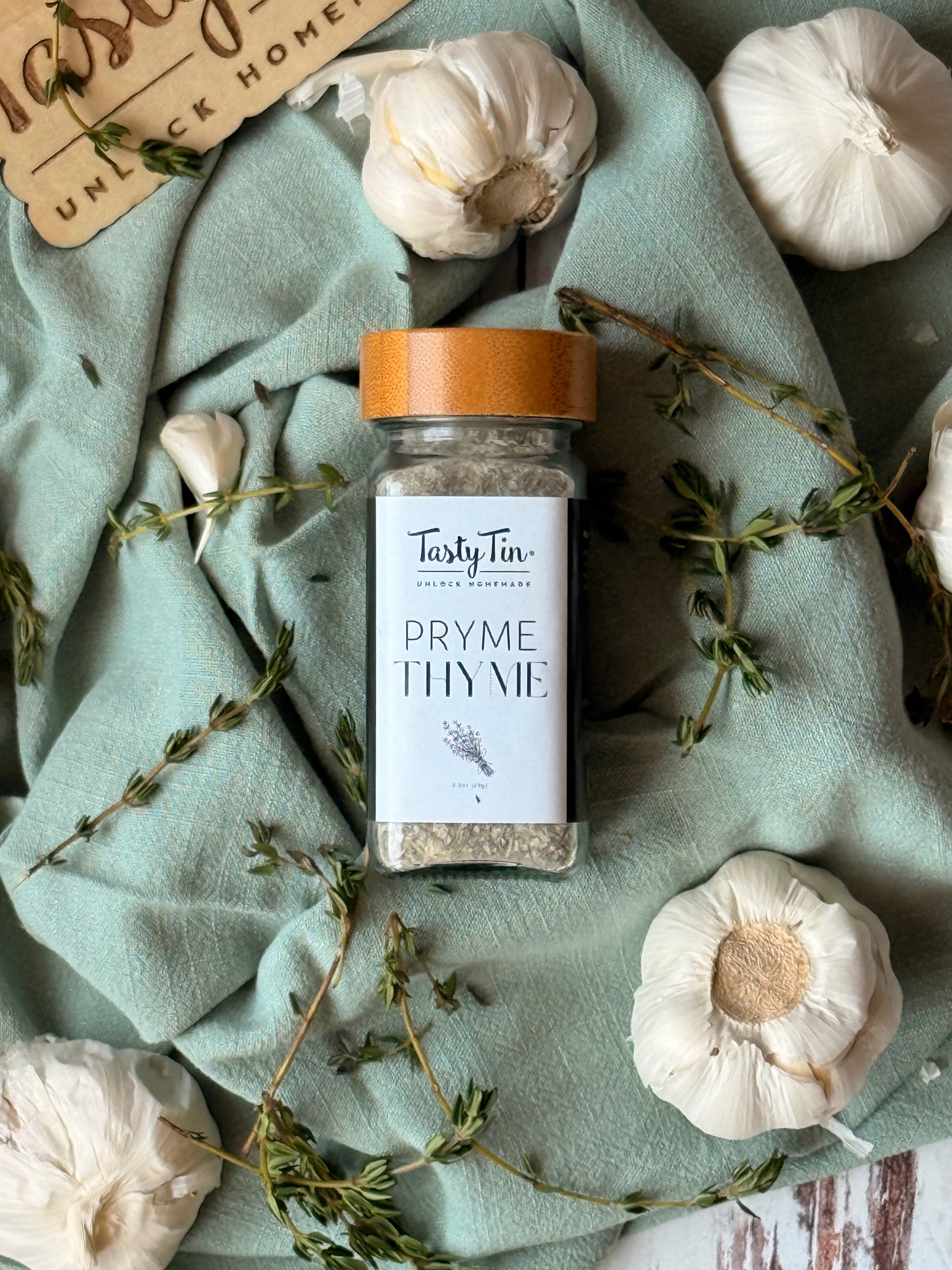 Tasty Tin thyme seasoning shaker with wooden lid surrounded by garlic and fresh thyme on green cloth