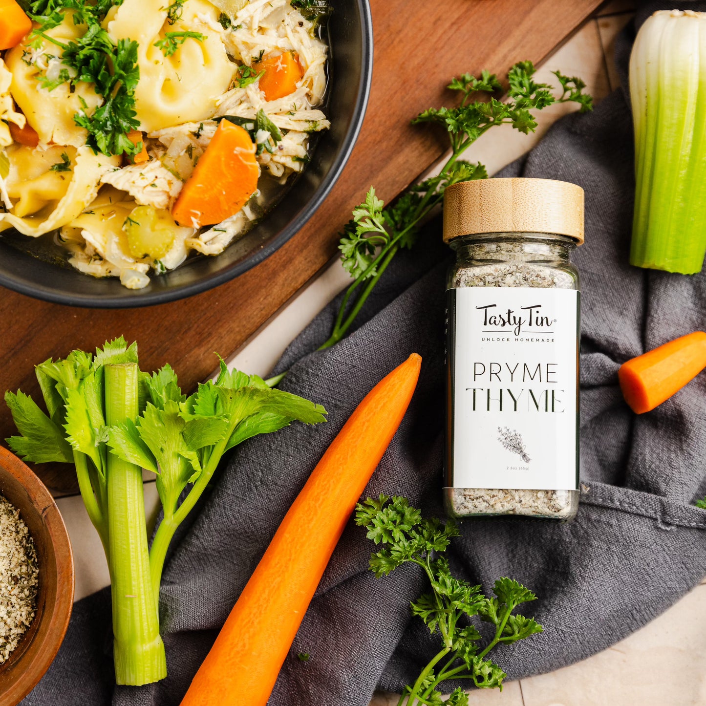 Tasty Tin thyme seasoning shaker with wooden lid surrounded by fresh vegetables and cooked dish